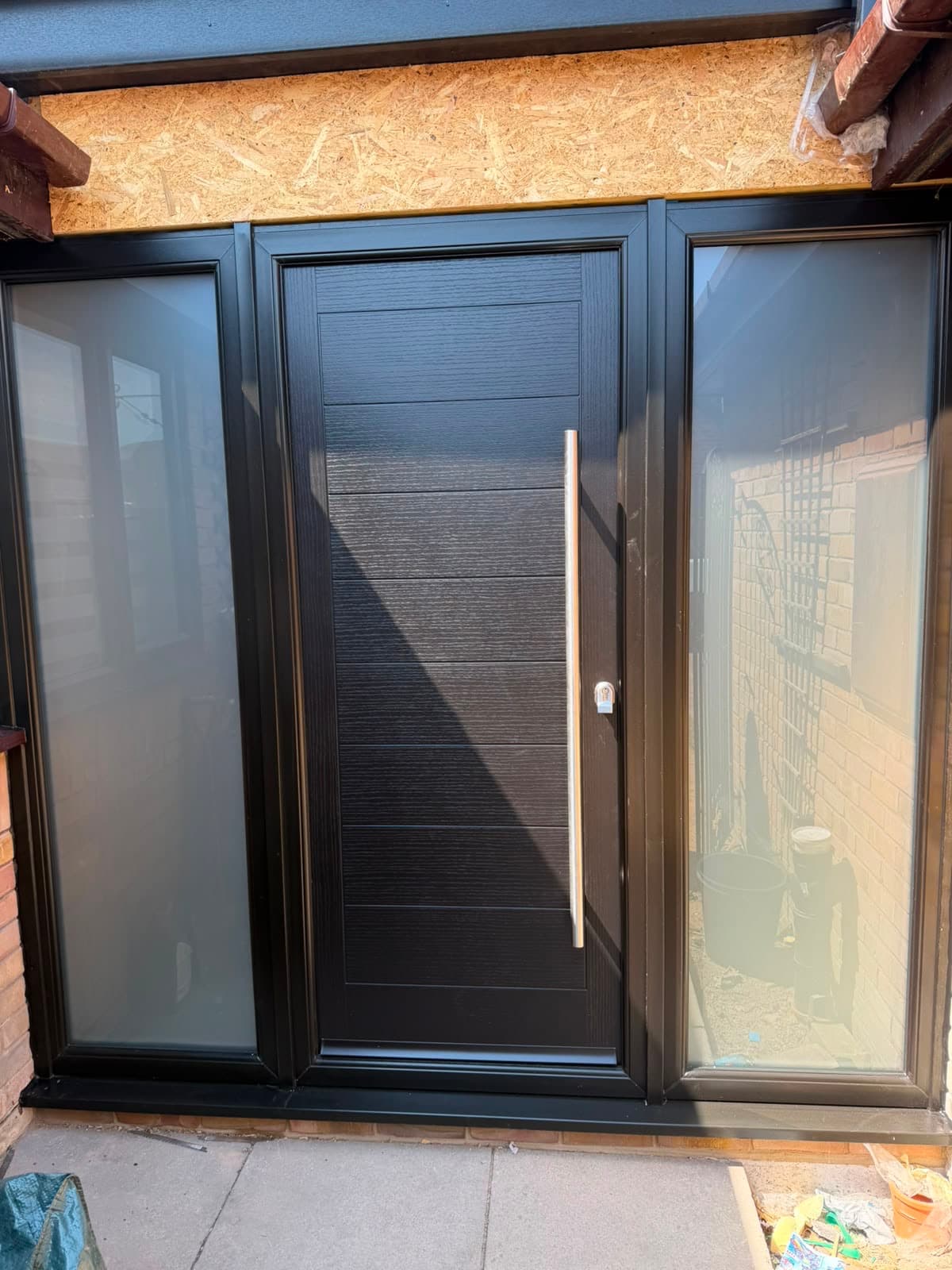 A dark woodgrain-finish composite front door with a narrow glazed panel and full-height sidelights, at a property still being finished